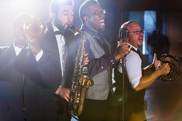 A jazz band performing at a nightclub.  The four male musicians are standing together, playing a trumpet, saxophone, and tambourine.  A mature, African American man is the lead singer, singing into a microphone.